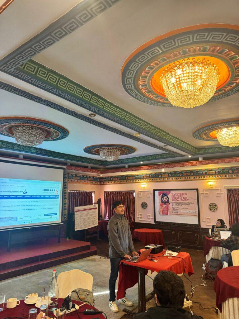 A person stands at a podium in a conference room with ornate ceilings, speaking to an audience. A large screen displays a website, and posters are visible in the background.
