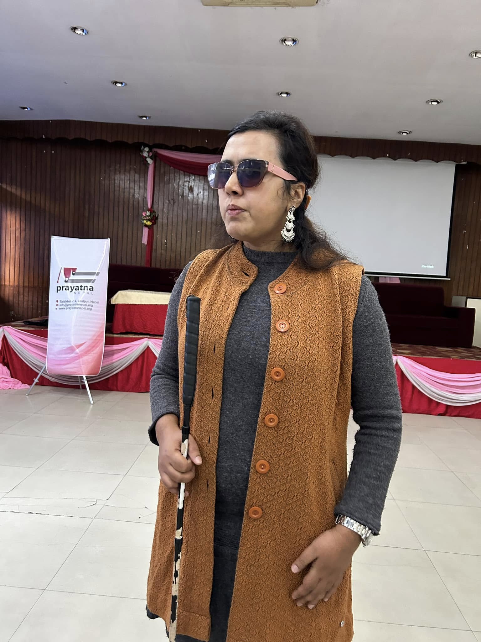 A woman stands in a brightly lit hall, holding a white cane, dressed in a brown vest over a gray sweater, with a stage in the background.
