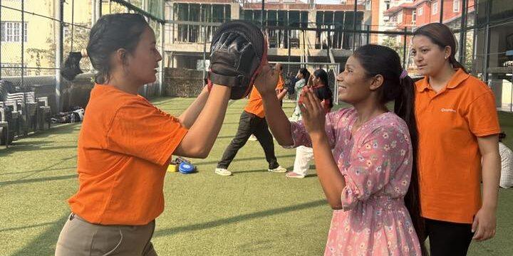 Two girls participate in a practice session, one catching with a glove while the other prepares to throw. The setting is outdoor sports facility.