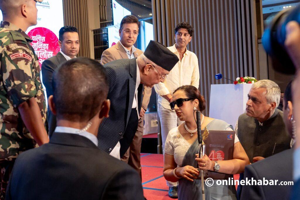 arita Lamichhane, wearing sunglasses and holding a certificate, is engaged in a conversation with the Prime Minister of Nepal (wearing a Dhaka Topi and grey suit). They are surrounded by other attendees, some looking on, at what appears to be a post-event gathering. Cameras and people are visible in the foreground, suggesting media presence.