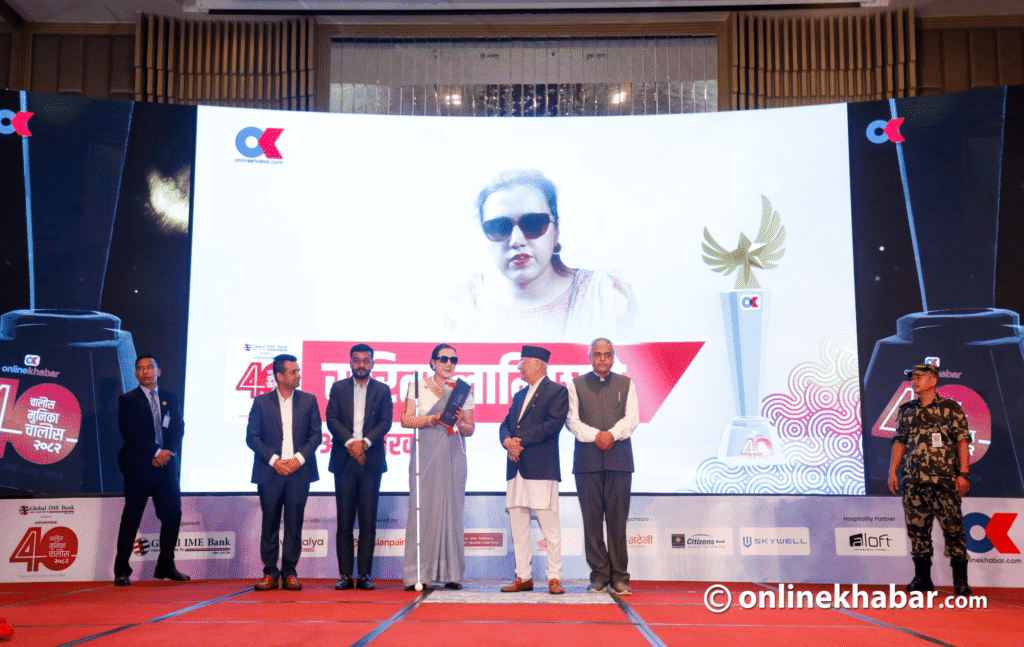 Sarita Lamichhane, a visually impaired woman in a grey saree and sunglasses, stands on a red carpeted stage holding a certificate and a white cane. She is flanked by four men in suits and a man in traditional Nepali attire (Daura-Suruwal and Dhaka Topi). A large screen behind them displays an image of Sarita, the "Online Khabar" logo, and the "40 Under 40" branding.