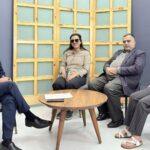 Four people sit in a semi-circle on chairs around a wooden coffee table in a studio setting with a light wooden grid backdrop. Two people are wearing sunglasses.