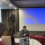 A man in a wheelchair holds a microphone and speaks to a seated woman in a conference room with a blank projection screen in the background.