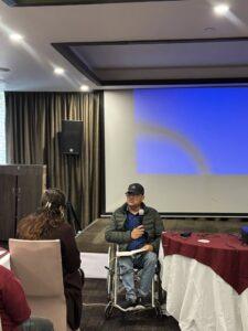 A man in a wheelchair holds a microphone and speaks to a seated woman in a conference room with a blank projection screen in the background.