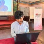 A participant in a light blue shirt is seated at a table, focused on a laptop during a training session. Two microphones lie on the red tablecloth in the foreground, and the background includes a projector screen, a presentation area, a whiteboard, and a Prayatna Nepal banner in a conference hall.