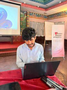 A participant in a light blue shirt is seated at a table, focused on a laptop during a training session. Two microphones lie on the red tablecloth in the foreground, and the background includes a projector screen, a presentation area, a whiteboard, and a Prayatna Nepal banner in a conference hall.