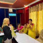 A woman presents to a group of seated women in a colorful room; one participant in yellow stands and gestures while others watch and smile.