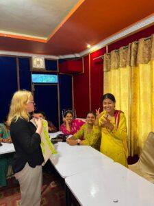A woman presents to a group of seated women in a colorful room; one participant in yellow stands and gestures while others watch and smile.