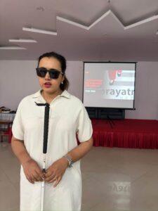 Ms. Sarita Lamichhane seen speaking as she is clad in white long dress and holding white cane in between her body with both hands, standing in-front of big hall where Prayatna Nepal’s banner being displayed in Projector screen behind her