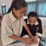 A blind girl participant is standing and holding a pen while touching her friends hand who is seated in a table-chair and are attempting to write alphabet in braille paper