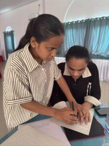 A blind girl participant is standing and holding a pen while touching her friends hand who is seated in a table-chair and are attempting to write alphabet in braille paper